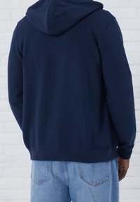 Uomo che indossa una felpa blu navy e jeans azzurri, mostrato da dietro contro uno sfondo di muro di mattoni bianchi.