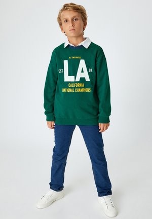 Sudadera verde con texto blanco y amarillo, jeans azules y zapatillas blancas. La sudadera presenta "LA" y "CAMPEONES NACIONALES DE CALIFORNIA."
