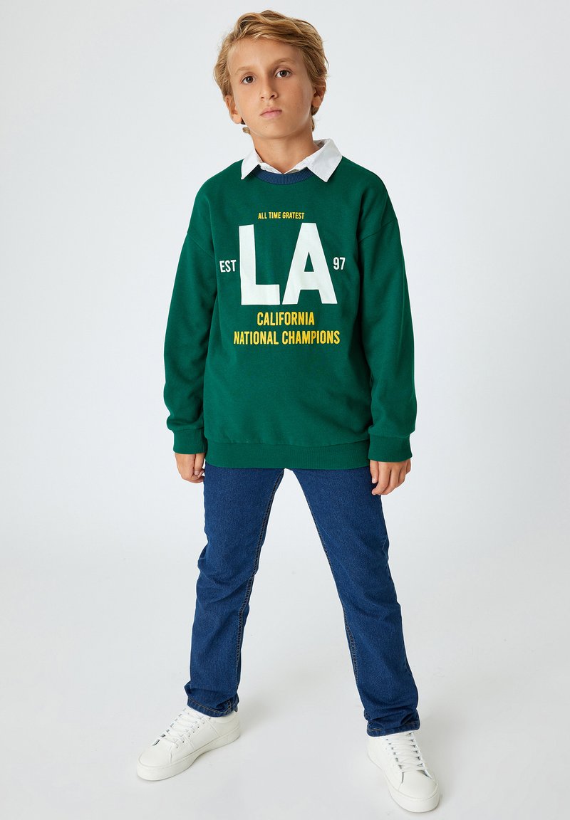 Zielona bluza z białym i żółtym napisem, niebieskie dżinsy i białe sneakersy. Bluza ma napis "LA" oraz "CALIFORNIA NATIONAL CHAMPIONS."