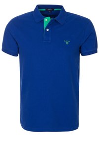 Blå polo skjorta i bomull, med en grön innerkrage och en liten grön logotyp på vänster sida. Korta ärmar och en klassisk passform.