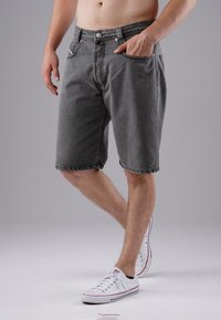 Man wearing gray denim shorts and white sneakers, standing with one hand in pocket against a plain gray background.