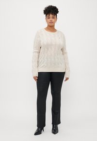 Pull crème en tricot torsadé avec des manches longues, un col rond et un détail logo subtil ; associé à un pantalon noir épuré et des bottines à bout pointu.