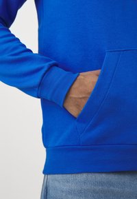 Niebieska bluza z kieszenią kangurzą, wykonana z miękkiego materiału. Posiada ściągacze przy mankietach i u dołu, o gładkiej fakturze i dopasowanym kroju.