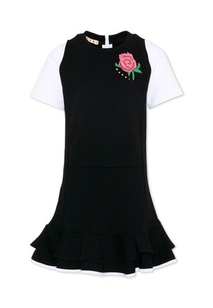 Zwarte mouwloze jurk gedragen over een wit shirt met korte mouwen, met roze rozenborduursel bij de schouder en een gerucheerde zoom.