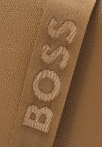 Brązowa skórzana torba z wyraźnym materiałowym paskiem z tłoczonym logo "BOSS". Widoczne są gładka tekstura i bogate detale kolorystyczne.