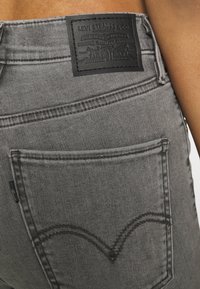 Šedé džíny z denimu s černou koženou nášivkou na pásu s raženým textem. Tmavé prošívání a zadní kapsa s charakteristickým designem.