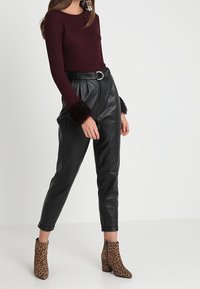 Vrouw in een bordeauxrode trui met bontmanchetten, zwarte leren broek met riem en laarzen met luipaardmotief, staande tegen een effen achtergrond.