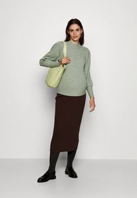 Nő zöld pulóverben, sötétbarna szoknyában, fekete cipőben, kezében világoszöld mintás válltáskával, egyszínű fehér háttér előtt áll.