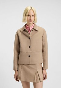 Beige wool blend jacket with black buttons, collared neckline, and two horizontal seams. Worn over a pink shirt and paired with a pleated skirt.
