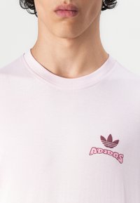 Helles rosa Baumwoll-T-Shirt mit Rundhalsausschnitt, versehen mit einem bedruckten dunkelroten Adidas-Logo und drei Streifenakzenten über dem Text.