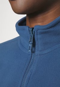 Niebieska kurtka polarowa z wysokim kołnierzem i przednim zamkiem błyskawicznym, cechująca się gładką teksturą i minimalistycznym designem.