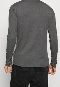 Eine Person trägt ein eng anliegendes, dunkelgraues Langarmshirt und schwarze Hosen, von hinten gezeigt vor einem schlichten, hellen Hintergrund.