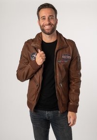 Braune Lederjacke mit Reißverschluss, gerippten Bündchen und Patches. Glatte Textur mit einem taillierten Design, kombiniert mit einem schwarzen Shirt darunter.