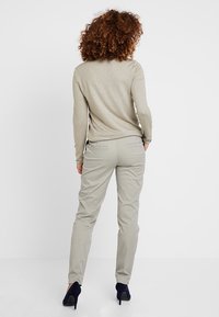 Licht beige langemouw top met een relaxte pasvorm, gecombineerd met lichtgrijze broek met achterzakken. Donkerblauwe hakken maken de outfit af.