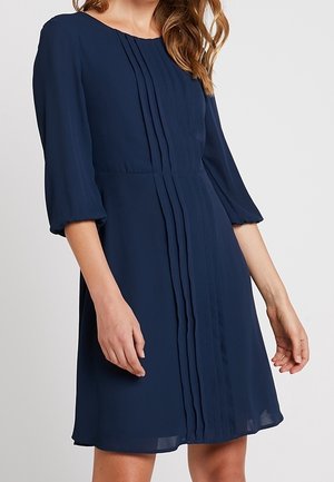 Marineblaues Kleid mit dreiviertellangen Puffärmeln, vertikalen Falten vorne und lockerem Schnitt, gefertigt aus leichtem Stoff.