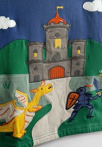 Tessuto di cotone ricamato che presenta un castello, un drago giallo e un cavaliere. Colori blu, verde, grigio e arancione con dettagli di cuciture e motivi elaborati.