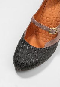 Chaussure Mary Jane noire texturée avec une bride en daim marron et une boucle dorée, dotée d'une semelle intérieure orange matelassée ornée de motifs circulaires.
