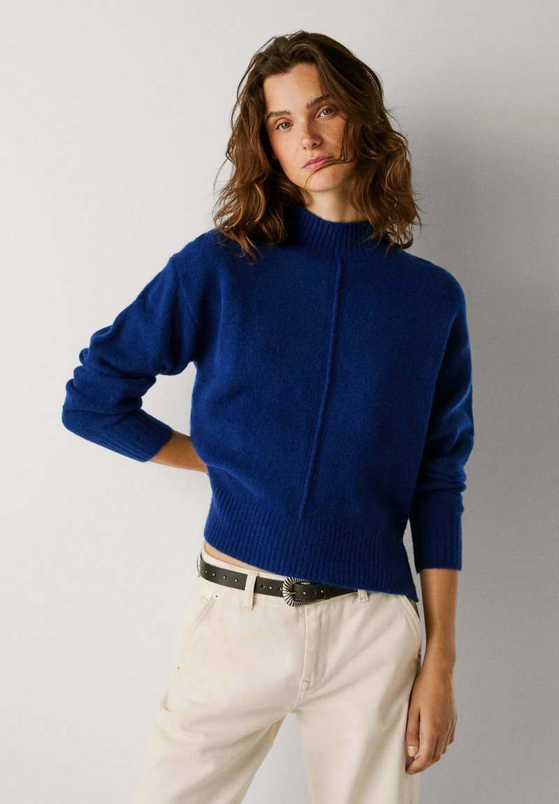 Modrý pletený svetr s vysokým límcem, žebrovanými manžetami a lemem; má středový šev jako ozdobný prvek. Kombinován s béžovými kalhotami a černým opaskem.