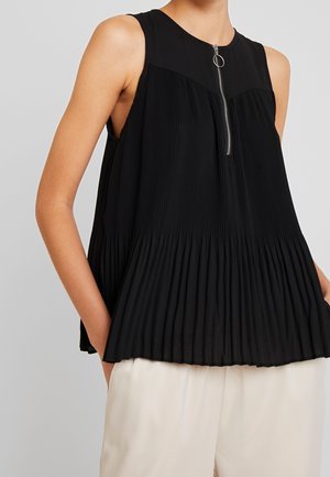 Ærmeløs sort plisseret top med en sølvfarvet lynlås foran og rund træk, båret med lyse beige bukser, hænderne i lommerne.