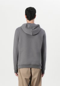 Sweatshirt cinza com capuz, com uma textura suave, punhos e bainha canelados, apresentando um corte descontraído. Usado sobre calças de cor clara.