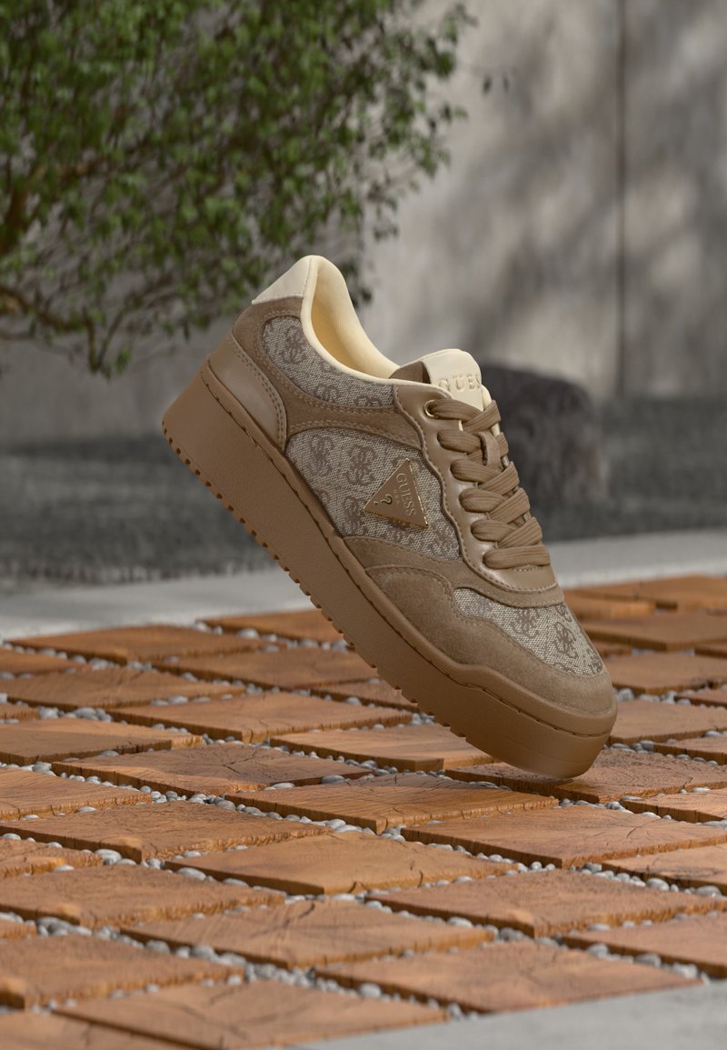 Μπεζ και καφέ αθλητικό παπούτσι Guess με μοτίβα λογότυπου και χοντρή σόλα που αιωρείται πάνω από μια ξύλινη και πλακόστρωτη υπαίθρια επιφάνεια.