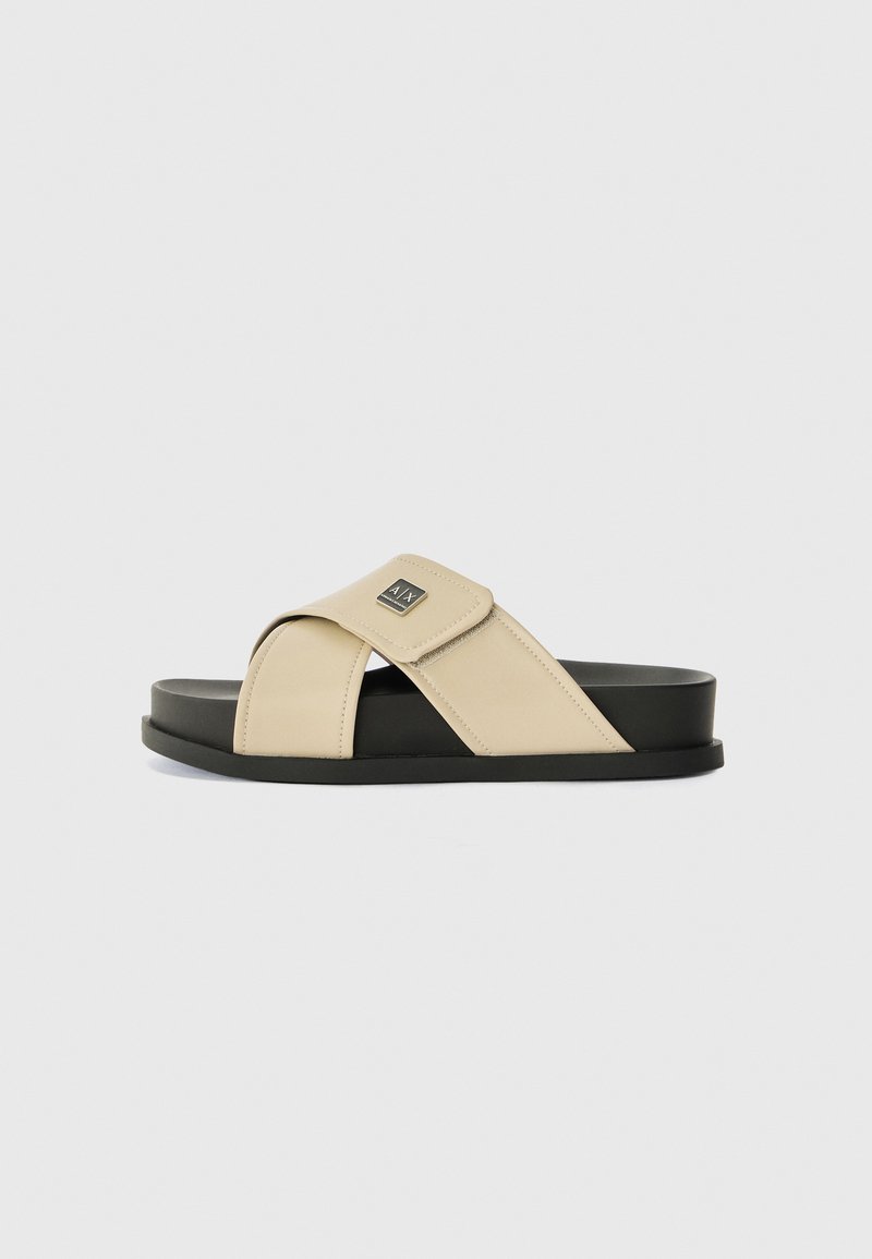 Beige szandál keresztbe tett pántokkal, sima anyagból készült, fekete gumi talppal. A pánton logo található. Minimalista dizájn.