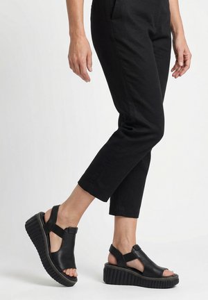 Osoba nosząca czarne krótkie spodnie i czarne sandały na platformie z odkrytymi palcami i elastycznymi paskami na białym tle.