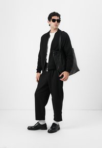 Chaqueta bomber negra sobre una camisa blanca, pantalones anchos negros, zapatos negros y gafas de sol. Sosteniendo un bolso de cuero negro.