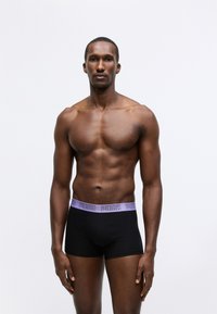 Sorte boxer shorts med en lilla Puma-logo i linningen, tettsittende design og en glatt tekstur. Bært av en mannemodell med atletisk kropp.