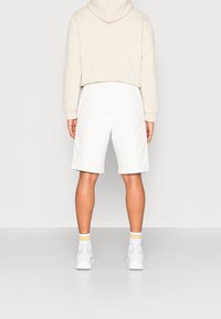 Shorts blancs à coupe décontractée, associés à un sweat à capuche crème. Baskets blanches visibles et chaussettes blanches à rayures. Design simple et minimaliste.