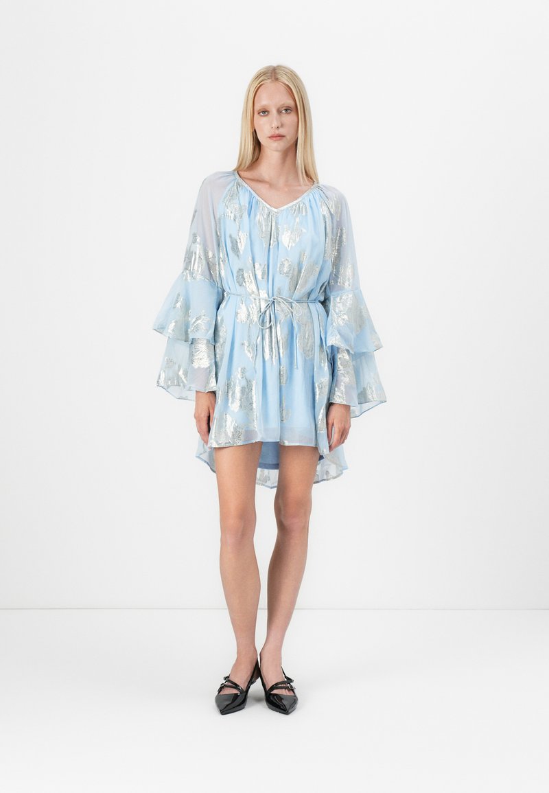 Vestido azul claro com um padrão floral em prata, mangas fluidas, cintura com cordão e barra assimétrica. Usado com sapatos apontados pretos.