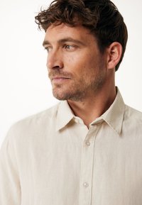 Camisa de lino beige con un collar de punta, botones sencillos y una tela texturizada, llevada por una persona con cabello corto rizado y piel clara.