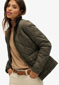 Geverfde olijf groene jas met een ritsluiting. Bevat een kraag, drukknoopsluiting en een bruine leren patch. Draag over een beige trui.