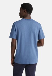 Muž stojící zády, oblečený v modrém tričku s krátkým rukávem a tmavých kalhotách na hladkém světlém pozadí.