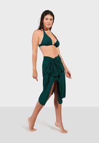 Tmavě zelený bikini s halter krkem a odpovídajícím zavinovacím pláštěm v sarongovém stylu, který má vysoký rozparek a zvlněné okraje.