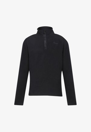 Młoda kobieta stojąca z rozluźnionymi rękami, ubrana w czarną bluzkę z długim rękawem i różowe spodnie w panterkę, na jednolitym białym tle.