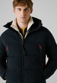 Chaqueta acolchada negra con capucha, que presenta un cuello de pelo sintético, bolsillos con cremallera y tiradores de cremallera en rojo contrastante. Textura suave y acolchada.