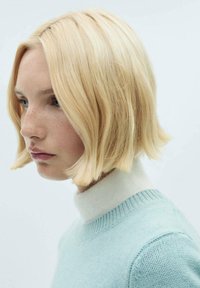 Kratka blond frizura v bob stilu. Oblači lahek modri pulover z rebrastim okroglim izrezom in belim ovratnikom. Gladek, teksturiran material.