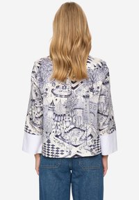 Loszittend shirt met een gedetailleerd blauw-wit patroon met speelse landschappen, bomen en architectuur; lange mouwen, witte manchetten.