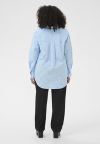 Chemise bleu clair à manches longues avec petits motifs de cœurs bleus, col boutonné et ourlet incurvé, associée à un pantalon noir ample.