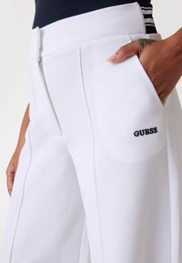 Vita vida byxor med vida ben gjorda av texturerat tyg, med en sidoficka och en svart "GUESS" logotyp på vänster lår. Subtila pressvecksdetaljer.