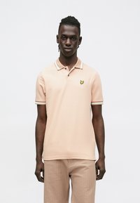 Világos rózsaszín galléros póló hegyes gallérral, három gombbal és sárga pillangó logóval a bal mellrészen. Bordázott mandzsetták fehér díszítéssel.