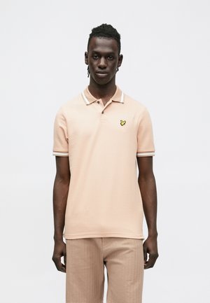 Hellrosa Polohemd mit spitzem Kragen, drei Knöpfen und gelbem Schmetterlingslogo auf der linken Brust. Gerippte Bündchen mit weißen Akzenten.