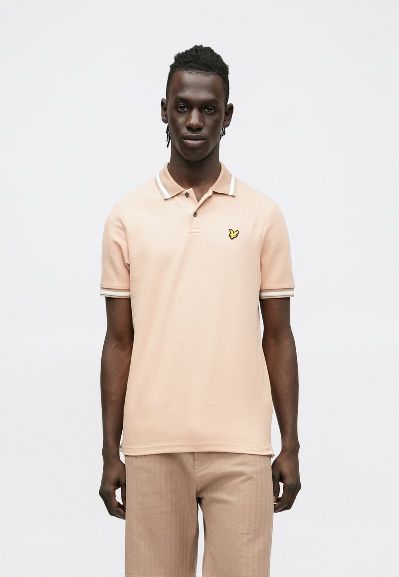 Világos rózsaszín galléros póló hegyes gallérral, három gombbal és sárga pillangó logóval a bal mellrészen. Bordázott mandzsetták fehér díszítéssel.