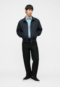 Νεαρός άνδρας στέκεται με τα χέρια στις τσέπες, φορώντας μαύρο μπουφάν με φερμουάρ, ανοιχτό μπλε πουκάμισο, μαύρο παντελόνι και μαύρα αθλητικά παπούτσια με λευκές σόλες.