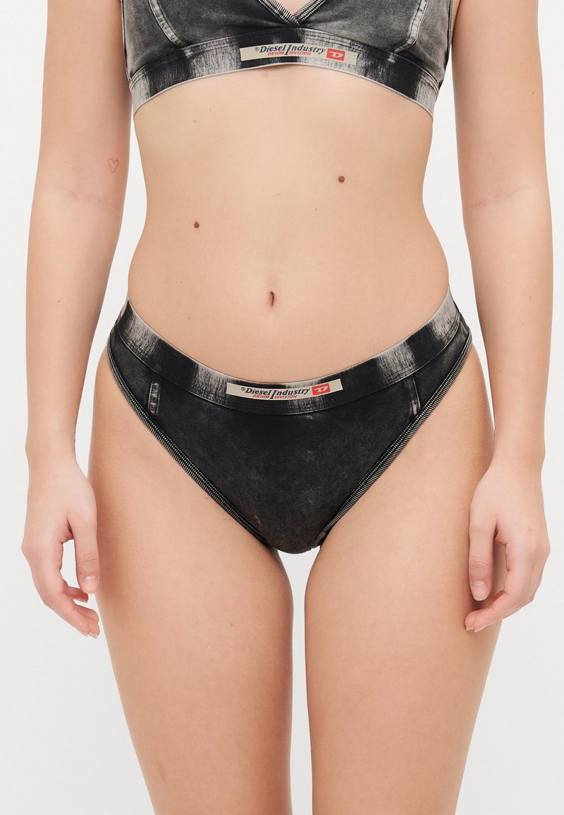 Svart denimstil bralette och högmidjade trosor med en slät textur. Har en logoband och dragkedjeaccentuationer på sidorna.