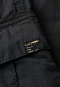 Zwart textiel met een subtiele textuur, met een duidelijk weergegeven zwart-gouden label waarop staat "SUPERDRY MFG CO. 076 CONTRACT."
