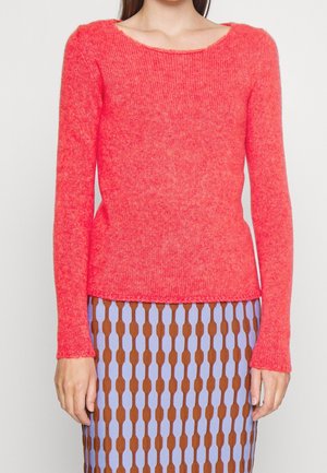 Femme portant un pull en maille à manches longues de couleur corail vif, associé à une jupe à motifs avec des formes ovales verticales marron et violet clair.