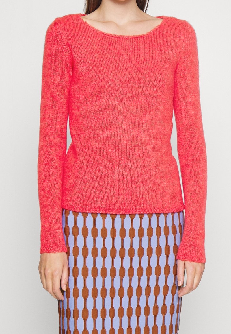 Femme portant un pull en maille à manches longues de couleur corail vif, associé à une jupe à motifs avec des formes ovales verticales marron et violet clair.