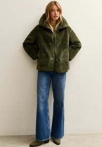 Kobieta w zielonej, puszystej kurtce z dużym kołnierzem, niebieskich dzwonowatych dżinsach i brązowych botkach za kostkę, stojąca przy jednolitej ścianie.